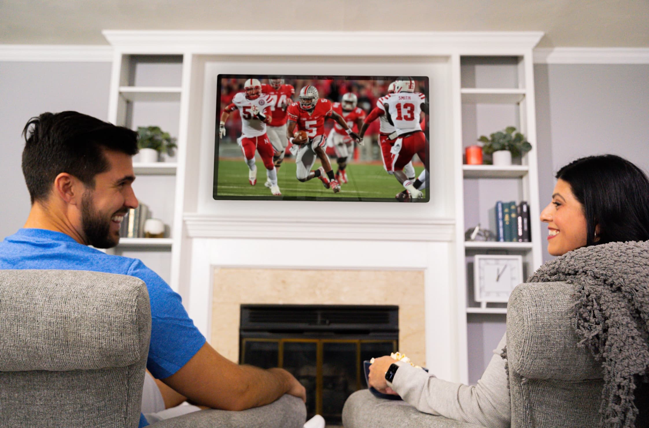roflect-blue-bg Couple watching football on a large screen above a fireplace. They are smiling and looking at each other while sitting in recliners.